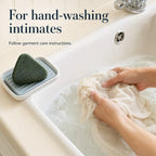 A practical hand-washing scene with a cream intimate garment in a utility sink and the original green triangular laundry soap beside the basin.